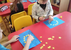 Dzieci układają 12 gwiazdek na wcześniej pomalowanym na niebiesko kartonie.